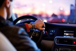 A man driving a car during sunset, focusing on his hand on the steering wheel and the dashboard, with a blurred cityscape in the background. He contemplates switching to the best car insurance for added