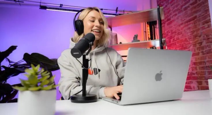 A woman smiles while recording a podcast, using a microphone and laptop in a studio with purple lighting and a brick wall background. SEO packages are visible on her laptop screen. A small plant is also on