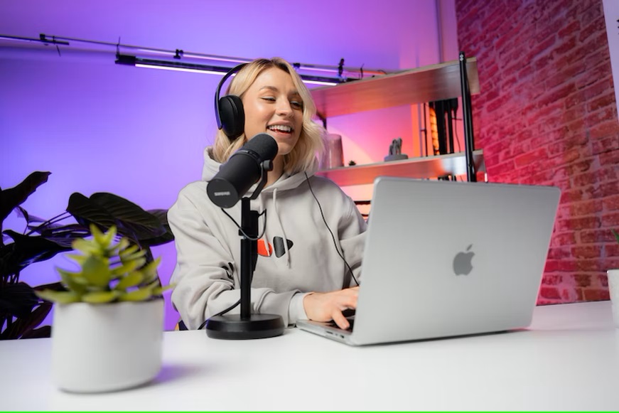 A woman smiles while recording a podcast, using a microphone and laptop in a studio with purple lighting and a brick wall background. SEO packages are visible on her laptop screen. A small plant is also on