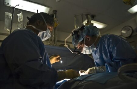 Two surgeons in scrubs and headlamps focus intently on a surgical procedure, benefiting from technological advancements, in a dimly lit operating room.