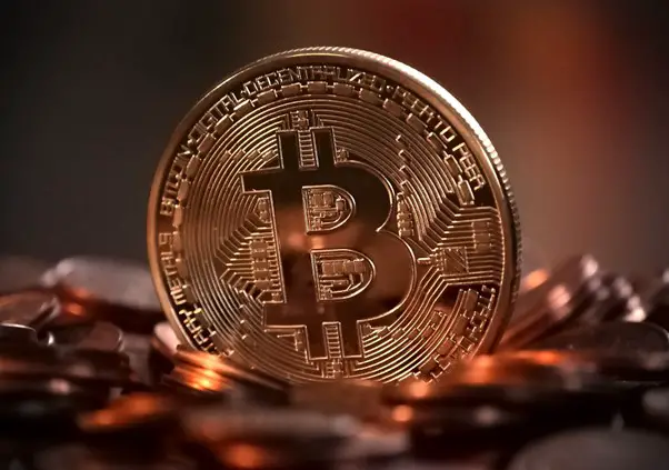 A close-up image of a shiny golden cryptocurrency coin standing upright on a pile of coins against a blurred red and gray background.