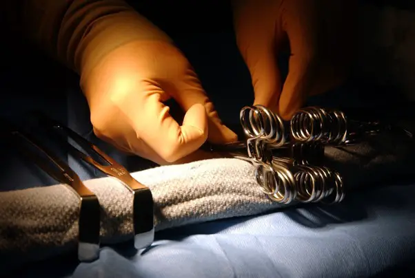 Close-up of a medical professional's gloved hands adjusting surgical instruments on a sterile blue cloth, illuminated by a focused light during a surgical procedure.