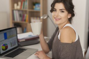 A young woman with shoulder-length dark hair sitting at a desk, smiling at the camera while holding a large white mug. She is working on a laptop displaying the Google Timeline and graphs on the screen,
