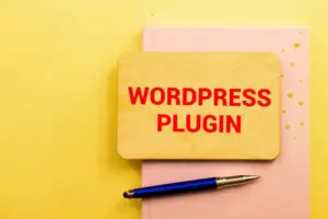 A wooden board with "Wordpress plugin detector" text in red, resting on a pink notebook with a blue pen, all against a yellow background.