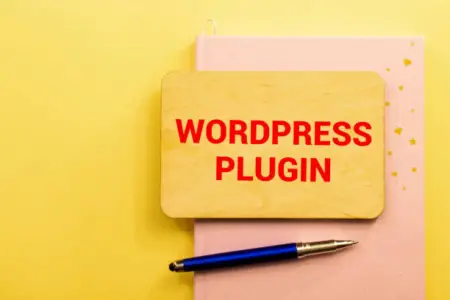 A wooden board with "Wordpress plugin detector" text in red, resting on a pink notebook with a blue pen, all against a yellow background.