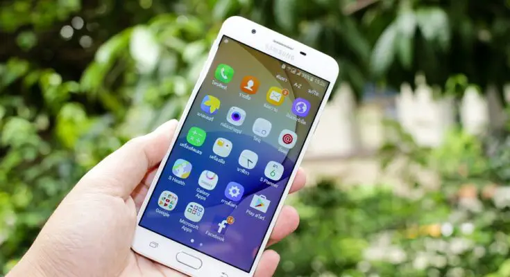 A hand holding a Samsung Android smartphone displaying colorful app icons on the screen, with a green leafy background.