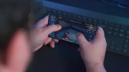 A person playing a shooter game on a mobile phone, focusing intently on the screen with their hands clutching the device over a keyboard in search of OSRS Gold. The dimly lit room adds