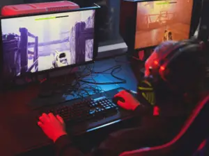 A person wearing headphones enjoys a gaming session, playing a first-person shooter video game on a computer with a vividly lit, large screen in a dark room.