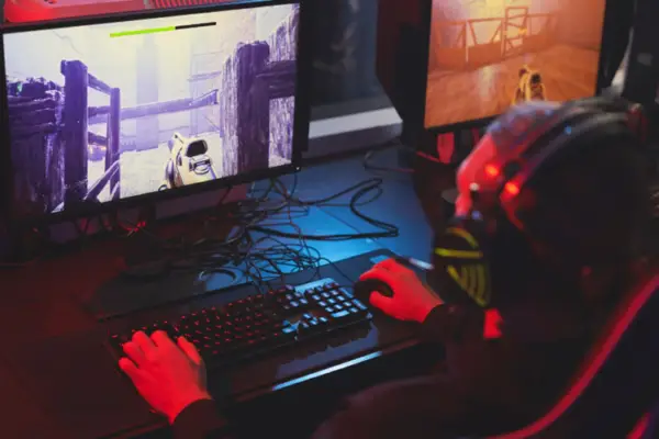A person wearing headphones enjoys a gaming session, playing a first-person shooter video game on a computer with a vividly lit, large screen in a dark room.