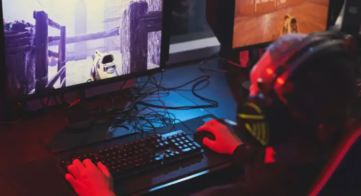 A person wearing headphones enjoys a gaming session, playing a first-person shooter video game on a computer with a vividly lit, large screen in a dark room.