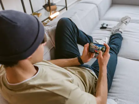 A young man wearing a beanie and t-shirt lounges on the floor, leaning against a sofa as he intently engages in gaming on his smartphone.