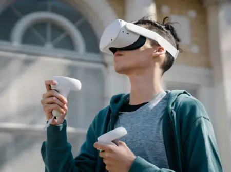 A young person engaged in outdoor gaming, wearing a VR headset and holding controllers, stands in front of a building with arched windows, seemingly interacting with a virtual environment.