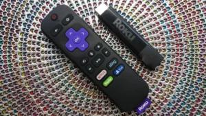 A Roku remote control lying next to a Roku Streaming Stick Plus on a textured, multicolored woven background.