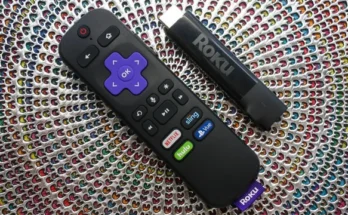 A Roku remote control lying next to a Roku Streaming Stick Plus on a textured, multicolored woven background.