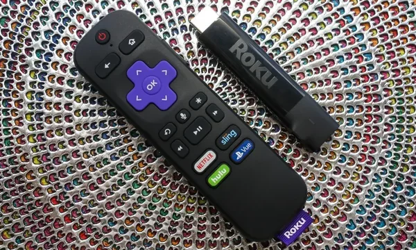 A Roku remote control lying next to a Roku Streaming Stick Plus on a textured, multicolored woven background.