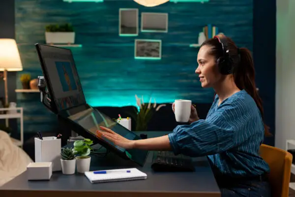 A woman wearing headphones works on multiple monitors in a cozy, well-lit home office with blue walls and decorative elements. She holds a coffee mug and is interacting with healthcare data on the screens.