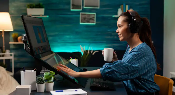 A woman wearing headphones works on multiple monitors in a cozy, well-lit home office with blue walls and decorative elements. She holds a coffee mug and is interacting with healthcare data on the screens.