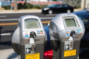 Two parking meters in focus with digital screens and credit card slots, standing on a city street with a blurred car parking and traffic in the background.