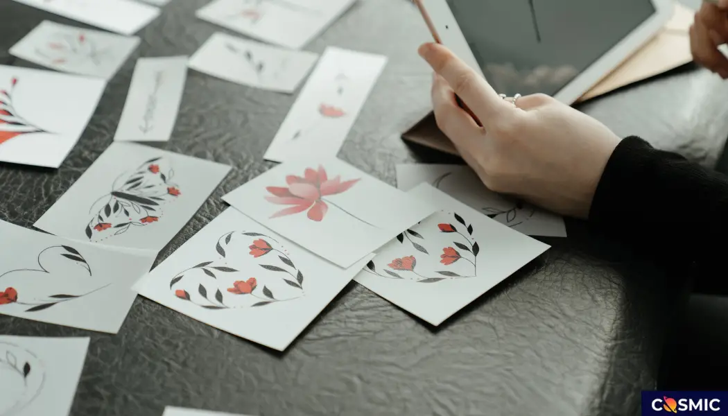 A person holding a stylus above a digital tablet, surrounded by various artistic floral cards laid out on a gray surface at Cosmicslot.