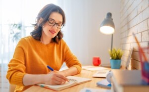 A woman in a yellow sweater and glasses writing about digital side hustles in a notebook at a desk with a laptop, lamp, and plant in a sunny room.