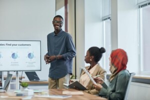 A smiling man stands presenting data analytics on a screen during a business meeting with two attentive female colleagues seated nearby.