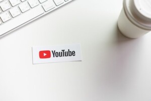 A YouTube logo sticker on a white desk, next to a computer keyboard and a takeaway coffee cup, symbolizing digital side hustles.