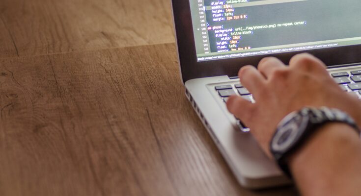 A person typing on a laptop with outsourced IT services code visible on the screen, placed on a wooden table. Focus on the person's wrist wearing a watch.