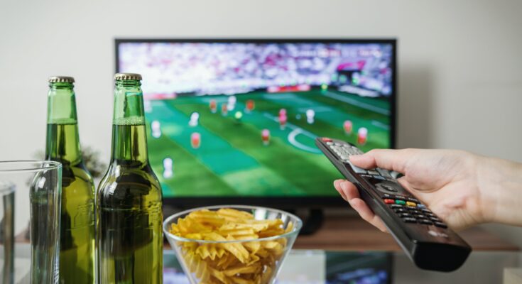 A person's hand holding a remote control, with beer bottles and a bowl of chips in front of a TV showing a football game and online betting odds.