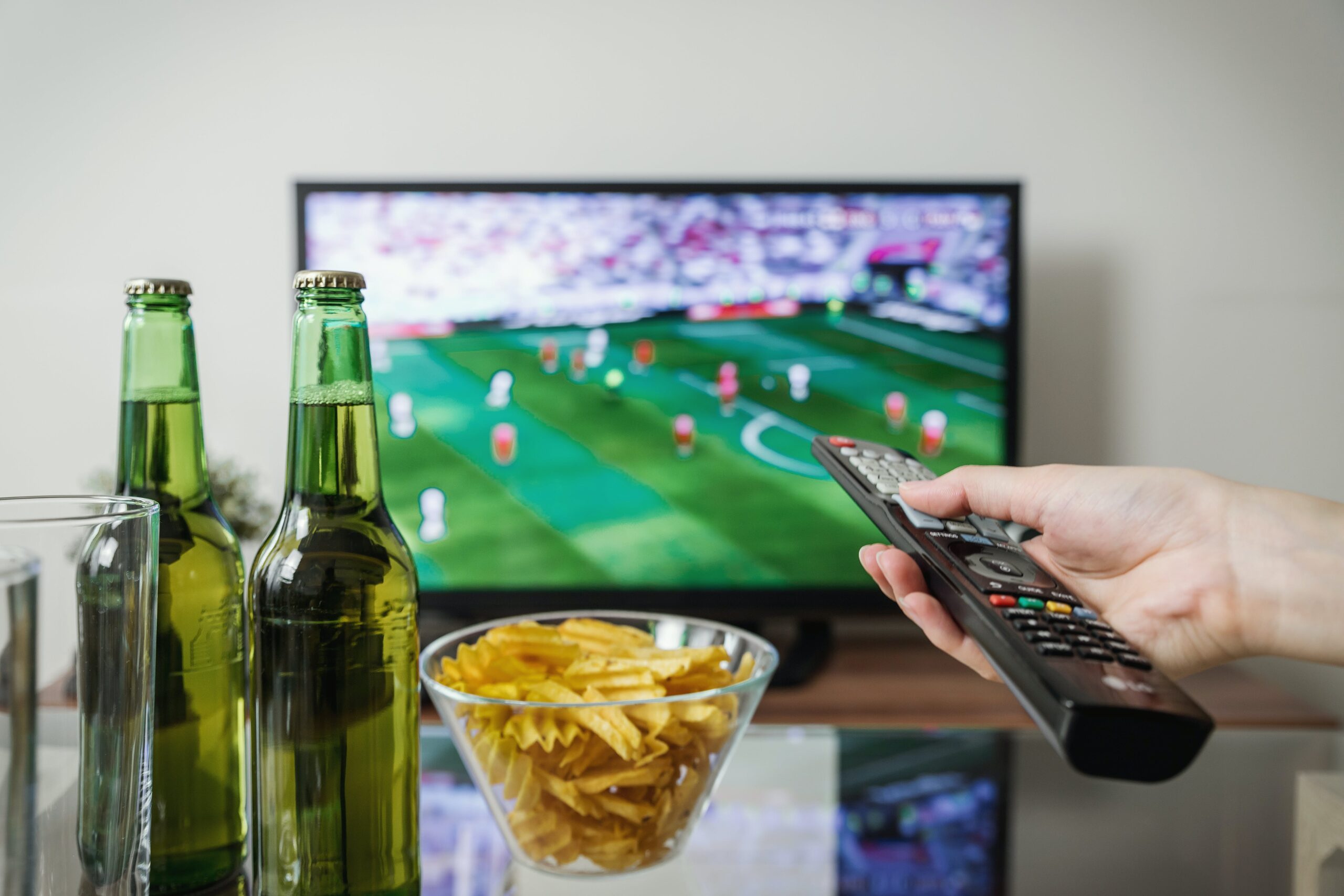 A person's hand holding a remote control, with beer bottles and a bowl of chips in front of a TV showing a football game and online betting odds.