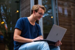 A young man with blond hair, wearing glasses and dressed casually in a blue t-shirt and jeans, smiling while working on outsourcing link building on a laptop outdoors.