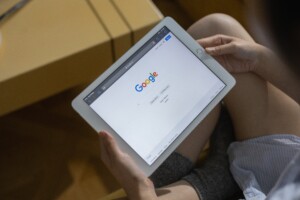 A person holding a tablet displaying the google search homepage, seated near a pile of moving boxes, suggesting a setting of organizing or outsourcing relocation.