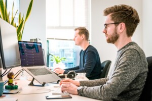 Two men working in an office, one focused on a computer screen with graphs for outsource link building, surrounded by tech gadgets, with another man blurred in the background. Bright, modern workspace.