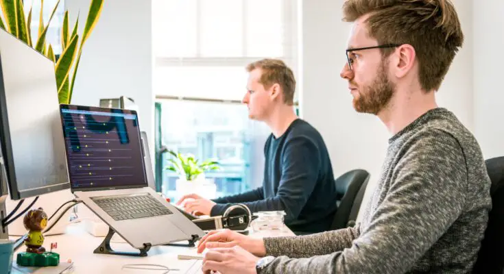 Two men working in an office, one focused on a computer screen with graphs for outsource link building, surrounded by tech gadgets, with another man blurred in the background. Bright, modern workspace.