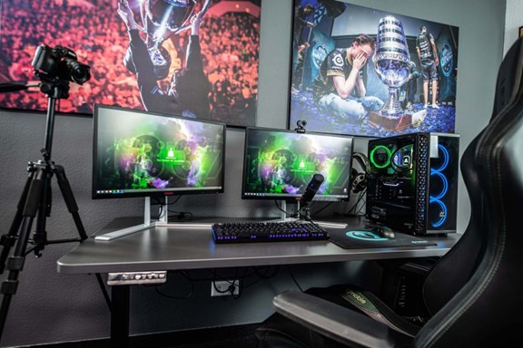 A modern gaming setup featuring three monitors displaying vibrant video game graphics, a DSLR camera on a tripod, and a desktop computer with LED lights, situated in a room with dim lighting and themed wall art