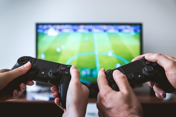 Two people holding gaming controllers while playing a soccer video game displayed on a screen, capturing their hands and part of the controllers.