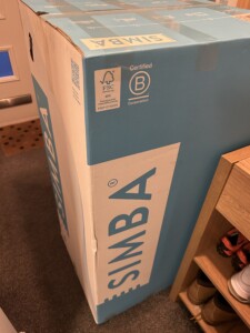 A large rectangular Simba mattress box in a room, leaning against a desk with visible shoes underneath and a chair in the background. The box is partially lit with soft lighting.