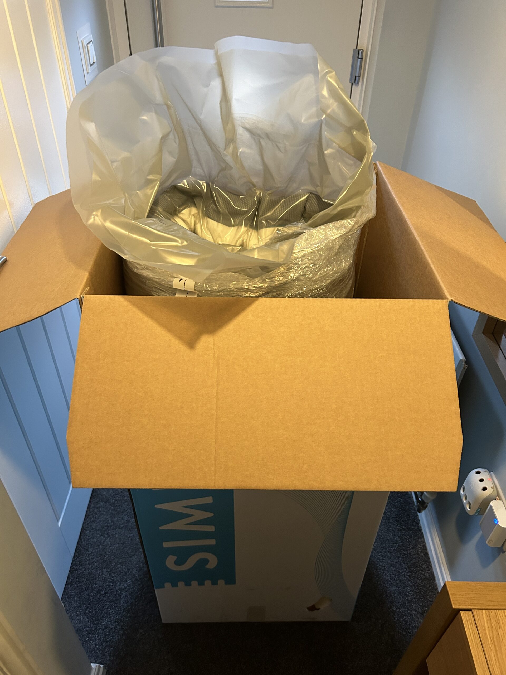 A large shipping box containing a Simba mattress stands open, revealing a thick protective plastic liner inside, set against a backdrop of a softly lit interior space.