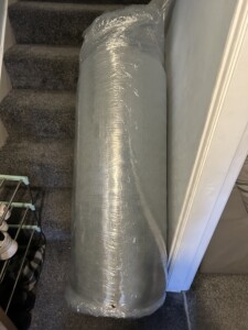 A large, cylindrical Simba mattress wrapped in clear plastic leaning against a grey carpeted staircase in a home, partially blocking the path.